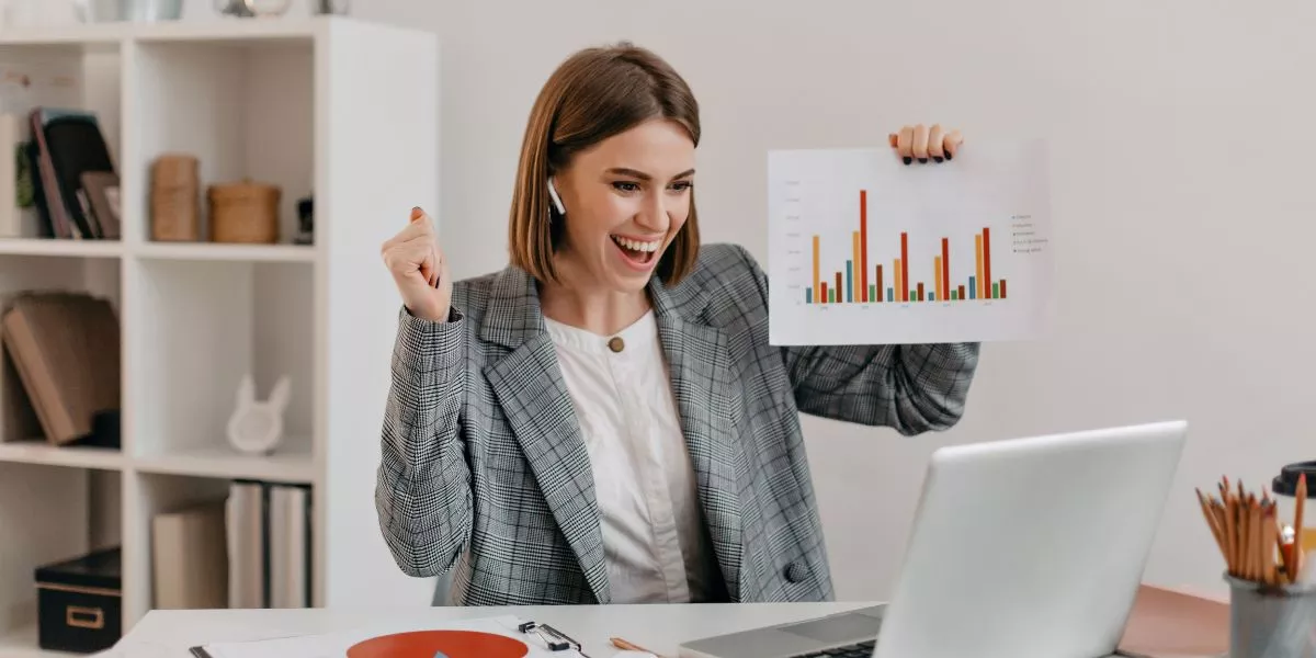 Mujer profesional celebrando resultados frente a su computadora en la oficina, mostrando gráficos de productividad laboral.